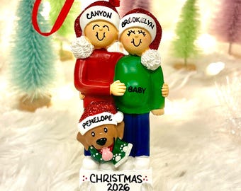 May include: A Christmas ornament featuring two figures and a dog wearing Santa hats. The hats are inscribed with the names Canyon, Brookelyn, and Penelope. The green shirt says BABY, and the base is inscribed with CHRISTMAS 2026.