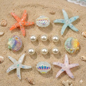 May include: A collection of beach-themed decorative items, including orange, blue, and pink starfish, iridescent seashells, and white pearls. The items are arranged on a sandy surface, evoking a coastal theme.
