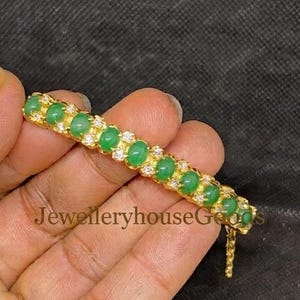 May include: A gold bracelet featuring a row of round, smooth, green cabochon stones alternating with small, clear, sparkling stones. The piece has a metallic yellow finish and a delicate chain link segment visible at the end of the main setting.