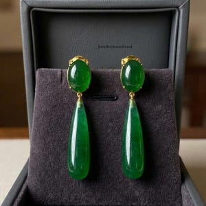 Op de afbeelding: Een paar goudkleurige oorbellen met ovale, jadegroene cabochons aan de bovenkant en langwerpige, druppelvormige groene hangers eronder. De oorbellen worden gepresenteerd in een met grijs fluweel bekleed sieradendoosje.