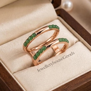 May include: Two rose gold rings featuring a row of small, round, green jade-like beads embedded along the top surface of each band. The rings are displayed together, and the text JewelleryhouseGoods is visible on the cream-colored fabric.