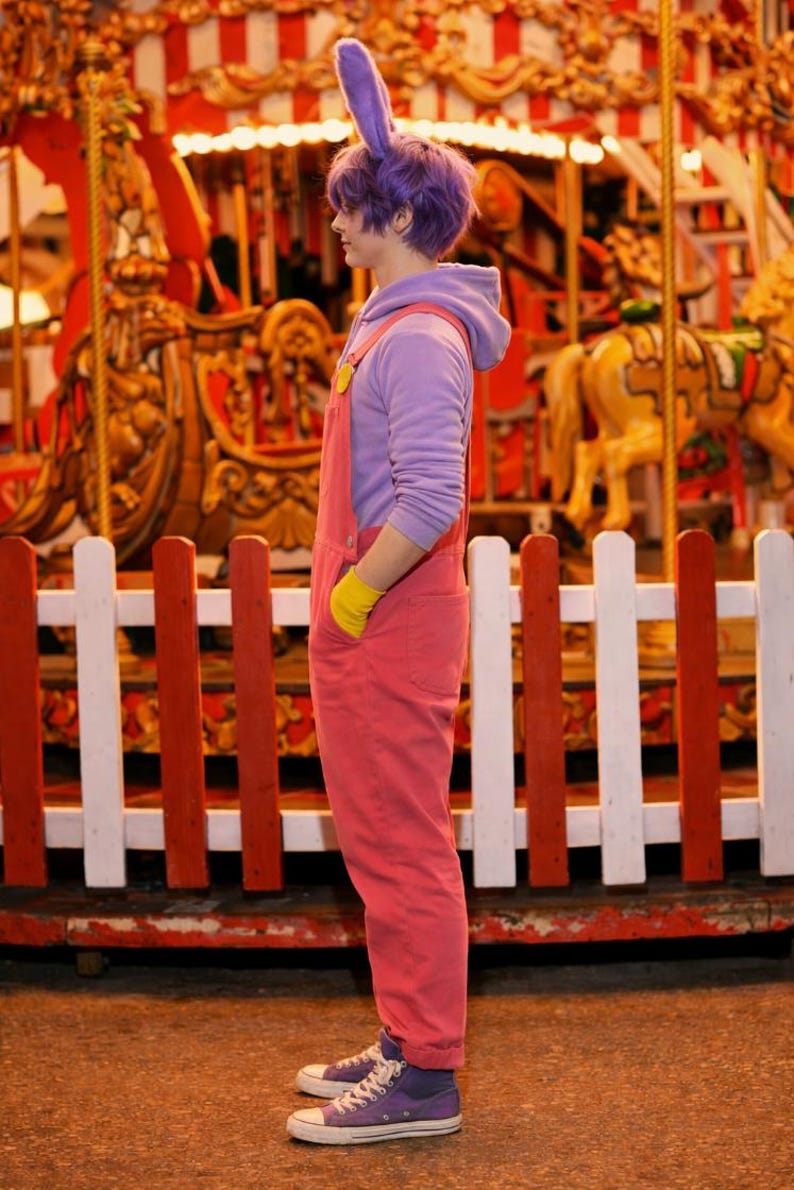 Pu&ograve; includere: Una felpa con cappuccio color lavanda indossata sotto una salopette di jeans rosa con guanti gialli e orecchie da coniglio viola. L'outfit include scarpe da ginnastica in tela viola con suola e punta in gomma bianca, cappuccio e tasche laterali.