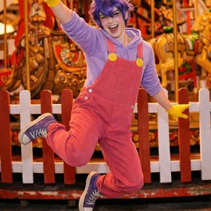 Pu&ograve; includere: Una persona indossa una felpa con cappuccio viola, una salopette rosa con bottoni gialli, guanti gialli e scarpe da ginnastica in tela viola. L'outfit include orecchie da coniglio viola su una parrucca viola, creando un costume da personaggio.