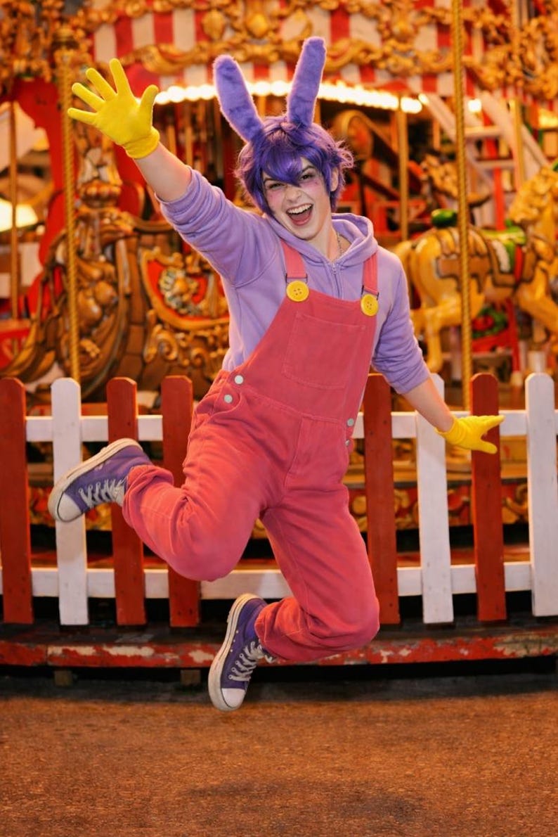 Pu&ograve; includere: Una persona indossa una felpa con cappuccio viola, una salopette rosa con bottoni gialli, guanti gialli e scarpe da ginnastica in tela viola. L'outfit include orecchie da coniglio viola su una parrucca viola, creando un costume da personaggio.