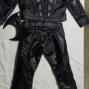 May include: A black leather costume set, including a jacket with padded shoulders and elbows, matching pants, and a mask. The jacket and pants have a quilted design. The mask is a stylized crescent shape.