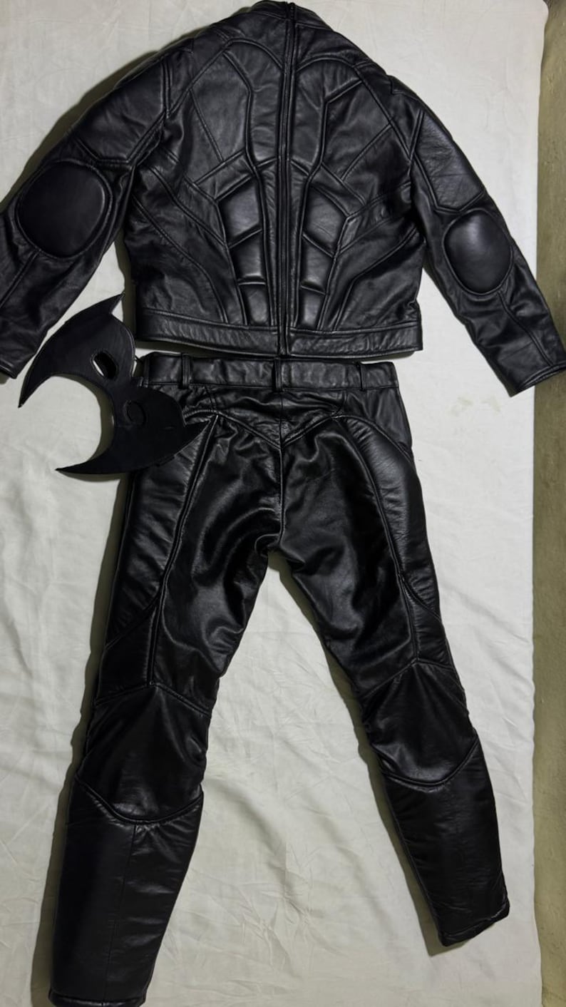 May include: A black leather costume set, including a jacket with padded shoulders and elbows, matching pants, and a mask. The jacket and pants have a quilted design. The mask is a stylized crescent shape.