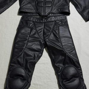 May include: Black leather motorcycle jacket and pants set. The jacket has a high collar and padded chest and shoulder details. The pants have knee pads and a tailored fit. The set is made of a shiny, durable leather material.
