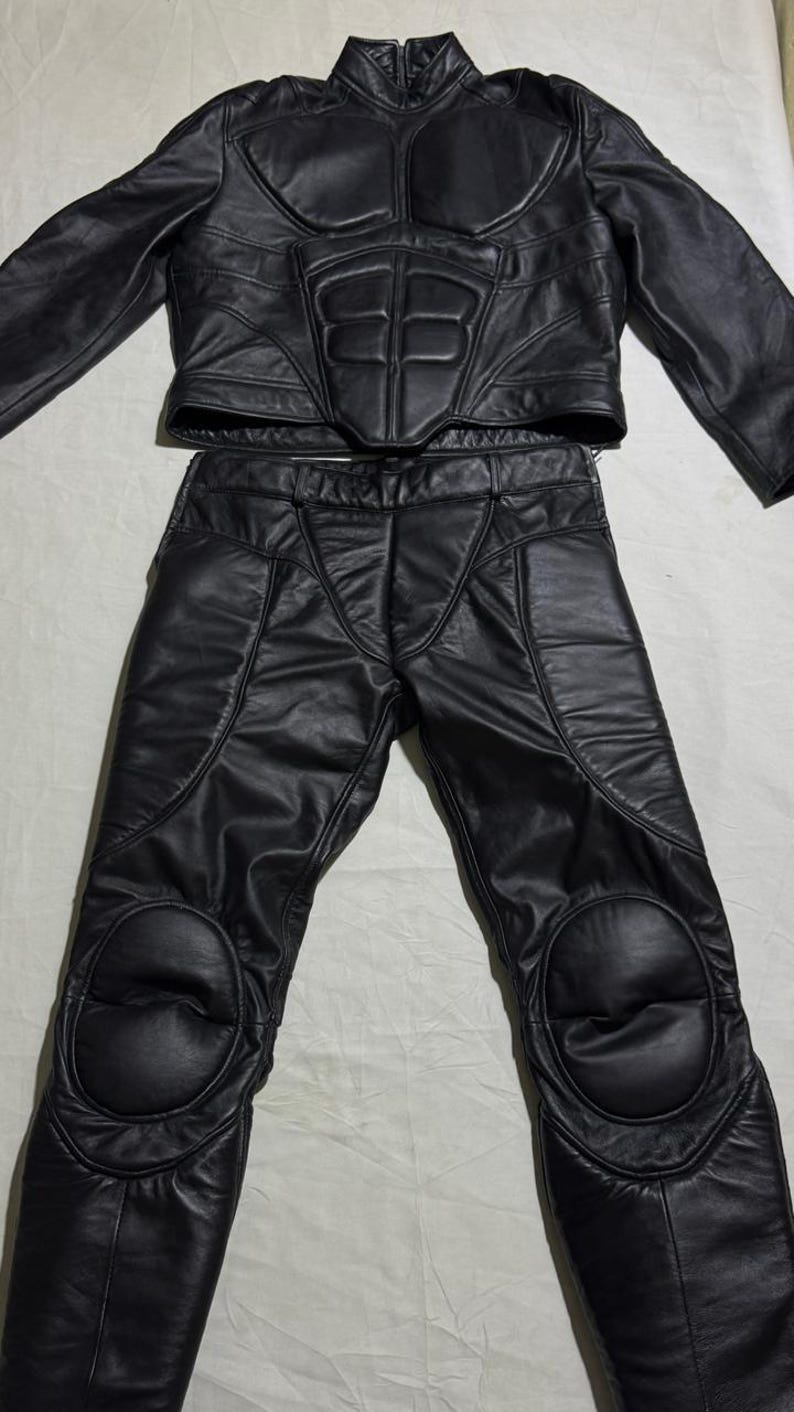 May include: Black leather motorcycle jacket and pants set. The jacket has a high collar and padded chest and shoulder details. The pants have knee pads and a tailored fit. The set is made of a shiny, durable leather material.
