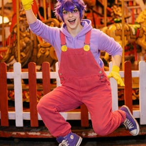 Pu&ograve; includere: Una persona indossa una felpa con cappuccio viola, una salopette rosa con bottoni gialli, guanti gialli e scarpe da ginnastica alte viola. L'outfit include una parrucca viola con lunghe orecchie da coniglio attaccate a un cerchietto.