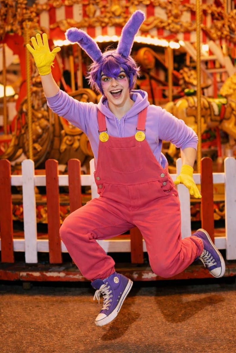 Pu&ograve; includere: Una persona indossa una felpa con cappuccio viola, una salopette rosa con bottoni gialli, guanti gialli e scarpe da ginnastica alte viola. L'outfit include una parrucca viola con lunghe orecchie da coniglio attaccate a un cerchietto.