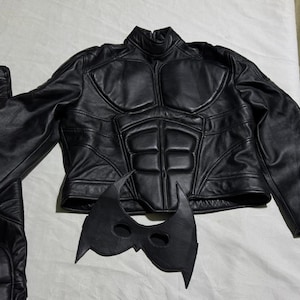 May include: A black costume set featuring a jacket, pants, and mask. The jacket has a padded design on the chest and shoulders. The mask is black with pointed edges. The pants are black.