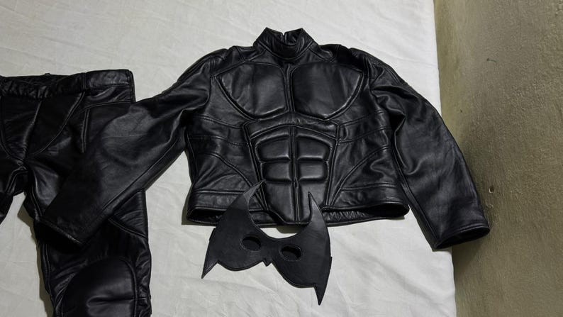 May include: A black costume set featuring a jacket, pants, and mask. The jacket has a padded design on the chest and shoulders. The mask is black with pointed edges. The pants are black.