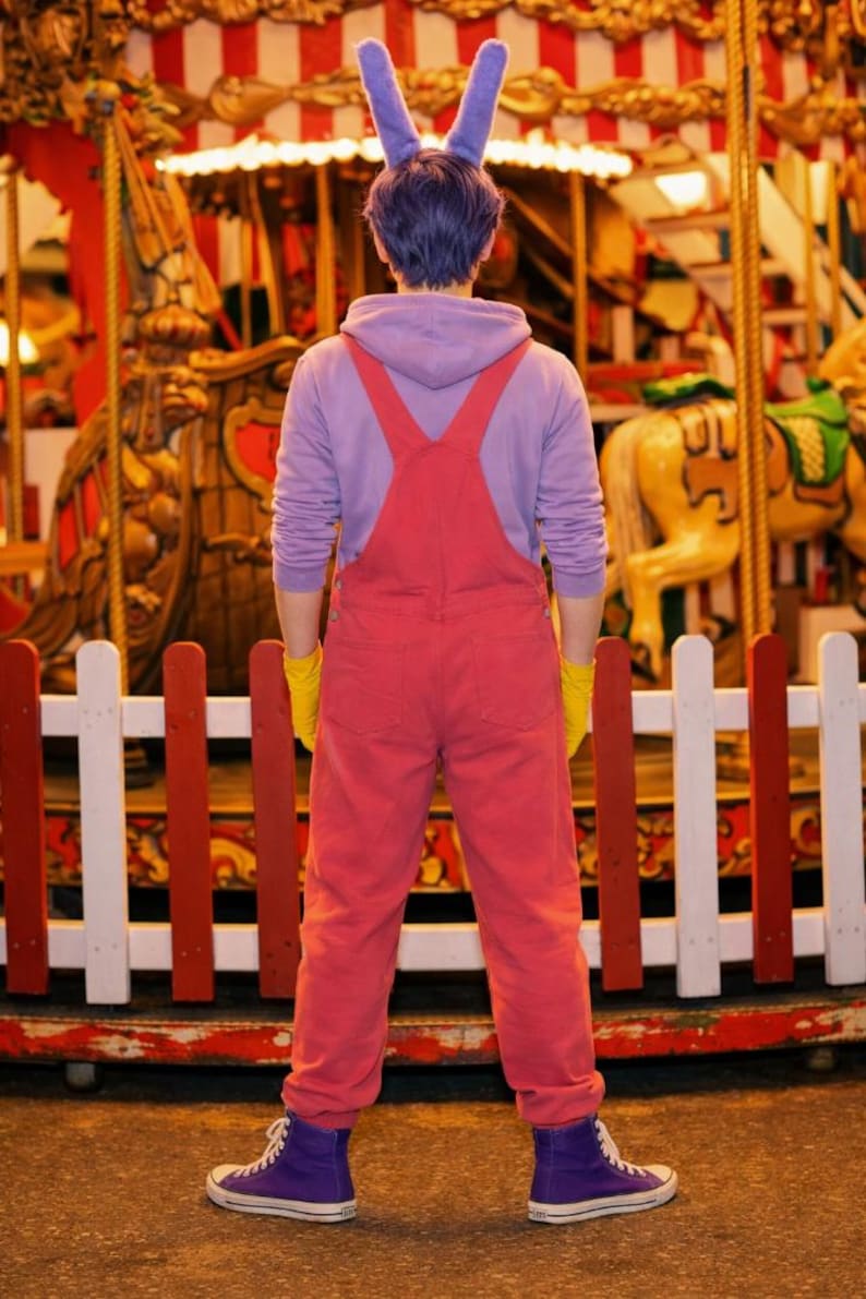 Pu&ograve; includere: Una felpa con cappuccio color lavanda indossata sotto una salopette di jeans rossa con guanti gialli, scarpe da ginnastica alte viola e una fascia viola con due orecchie da coniglio alte e dritte. L'outfit &egrave; visto di spalle.