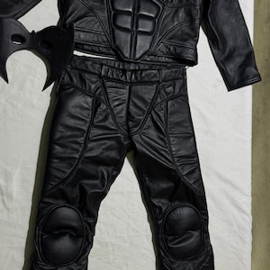 May include: Black leather costume set featuring a jacket, pants, and mask. The jacket has a high collar and padded chest and shoulder details. The pants have knee padding. The mask has a bat-like design. The costume is likely for cosplay or a themed event.