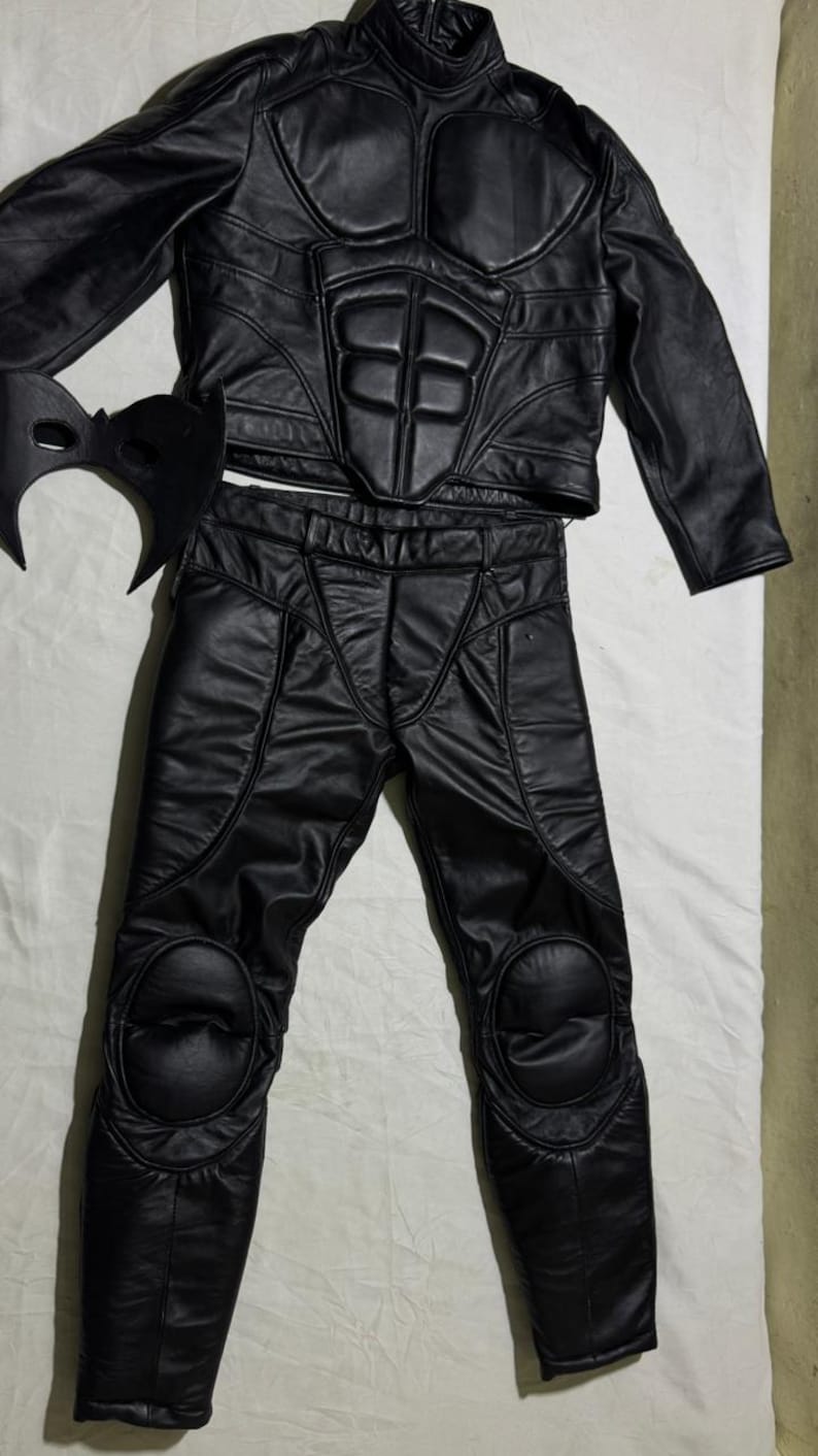 May include: Black leather costume set featuring a jacket, pants, and mask. The jacket has a high collar and padded chest and shoulder details. The pants have knee padding. The mask has a bat-like design. The costume is likely for cosplay or a themed event.