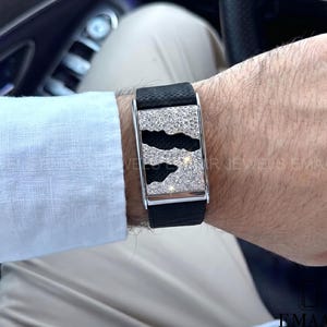 Puede incluir: Una pulsera de cuero negro con una hebilla rectangular plateada. La hebilla presenta un patrón de piedras claras y negras. La pulsera se lleva en la muñeca, con una manga de camisa blanca visible en el fondo.