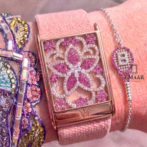 May include: A rectangular watch with a rose gold frame featuring a floral motif encrusted with pink and white stones on a pink fabric strap. Beside it is a silver chain bracelet with a tennis racket charm containing the letter B and pink stones.