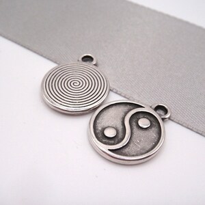 Könnte beinhalten: Zwei silberfarbene Anhänger. Einer zeigt ein Spiralmuster, der andere ein Yin-und-Yang-Symbol. Beide Anhänger haben oben eine kleine Schlaufe zum Befestigen. Sie liegen auf einem hellgrauen Band.