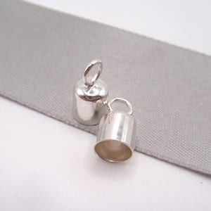 Könnte beinhalten: Zwei kleine, glänzende, silberfarbene Metallglocken mit einer Schlaufe oben. Eine steht aufrecht, die andere liegt auf der Seite. Sie liegen auf einem hellgrauen Stoff mit weißem Hintergrund. Es sind wahrscheinlich Schmuckzubehör.