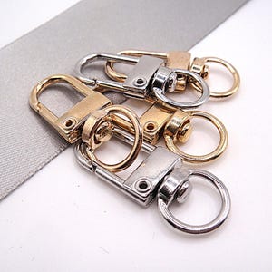 Könnte beinhalten: Eine Sammlung von drehbaren Karabinerverschlüssen aus Metall in Silber- und Goldtönen. Jeder Verschluss verfügt über einen rechteckigen Körper mit federbelastetem Schnapper und einen runden Ring an der drehbaren Basis zur Befestigung.