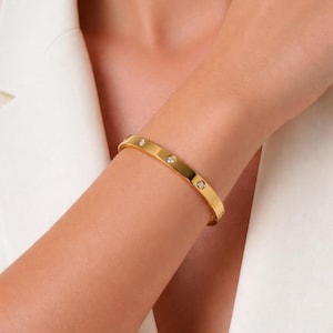 May include: A gold-coloured bangle bracelet with three clear, round gemstones. The bracelet is worn on a person's wrist. The background is a white blazer.