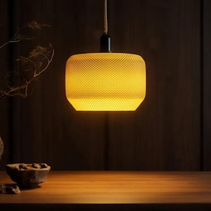 May include: A cylindrical pendant light with a textured, honeycomb-patterned yellow shade. The lamp emits a warm, glowing light from within, suspended by a simple cord attached to a black fixture, creating a focused illumination in the space.