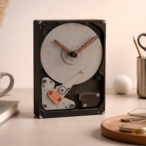 May include: A unique clock made from a repurposed hard drive. The clock face is a silver disc with orange hands and hour markers. The clock is set on a desk with a mug, book, and other office supplies.