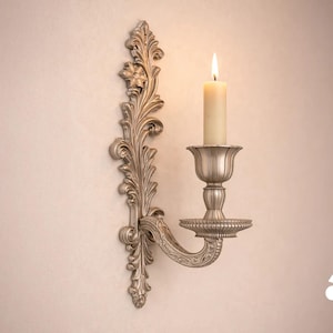 May include: A silver-toned wall sconce featuring an ornate, vertically elongated backplate with intricate leaf-like carvings. A curved arm extends outward to support a fluted candle cup holding a single, lit, cream-colored pillar candle.