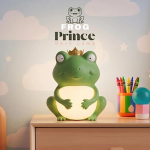 May include: A green frog-shaped desk lamp with a gold crown on its head and a glowing white belly. Above the lamp, the text reads FROG Prince Desk Lamp. The lamp is positioned on a wooden surface next to colorful pencil holders and a small sphere.
