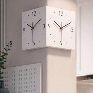 May include: Two white square clocks mounted on a wall. Each clock face has black numerals and hands, with gold-colored centers. The clocks are positioned at a right angle to each other. The background includes a wall, a decorative panel, and a glimpse of a room.