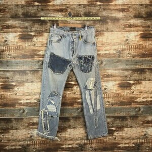 Può includere: Jeans in denim azzurro con chiusura a bottoni, diverse toppe usurate e dettagli in tessuto sfilacciato. I jeans presentano due tasche in denim blu scuro cucite sulle cosce anteriori e sono appesi a una gruccia in legno con un righello.