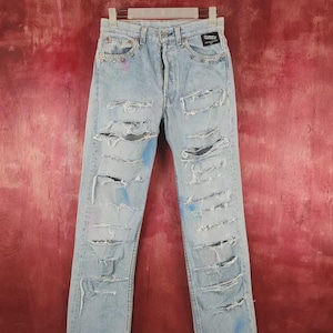 Può includere: Jeans in denim azzurro con ampie zone usurate, strappi e lacerazioni sulla parte anteriore delle gambe. I pantaloni presentano una chiusura a bottoni, rivetti in metallo e schizzi di vernice blu. Una piccola etichetta nera è cucita in vita.