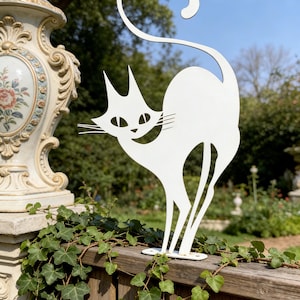 Könnte beinhalten: Eine weiße Metallskulptur in Form einer Katze mit gebogenem Schwanz, die auf einem Holzzaun steht. Die Katze hat ein lächelndes Gesicht und ist von Grün und einer dekorativen Steinsäule umgeben. Die Skulptur ist etwa 50 cm hoch.