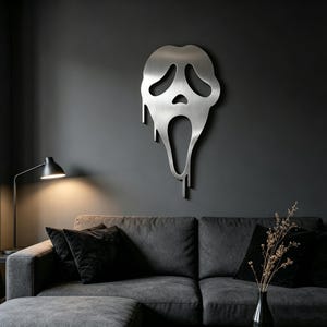 Puede incluir: Una pieza de arte mural de metal cepillado con la forma de la icónica máscara de Scream, con un efecto de goteo. La obra de arte está montada en una pared gris oscuro sobre un sofá seccional gris con cojines negros.