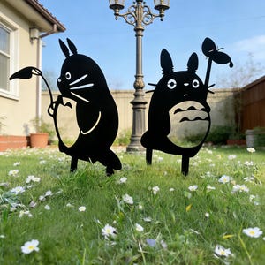 Könnte beinhalten: Zwei schwarze Metall-Gartendekorationen, die Zeichentrickfiguren darstellen. Eine hält ein Blatt, die andere einen Schmetterling. Sie stehen in einem grasbewachsenen Bereich mit weißen Blumen unter einer dekorativen Straßenlaterne.