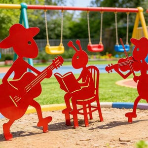 May include: Red metal cutouts of musical figures playing instruments in a playground setting. One figure plays a guitar, another reads a book, and a third plays a violin. Swings and playground equipment are in the background.