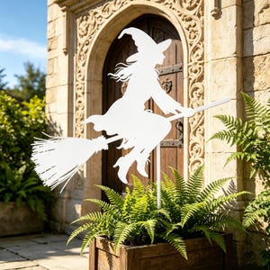 May include: A white metal silhouette of a witch riding a broomstick. The witch is wearing a pointed hat and flowing robes. The decoration is placed in a wooden planter box filled with green ferns, in front of a stone building with a wooden door.