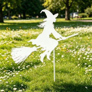 May include: A white metal silhouette of a witch riding a broomstick, set in a grassy field with wildflowers. The witch wears a pointed hat and flowing robes, with the broomstick extending horizontally. The artwork is mounted on a white stake.