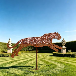 Op de afbeelding: Metalen sculptuur van een springende cheetah met een roestkleurige afwerking. De cheetah heeft een gedetailleerd vlekkenpatroon en is gemonteerd op een metalen paal. De sculptuur staat in een tuin met verzorgde gazons en beelden.