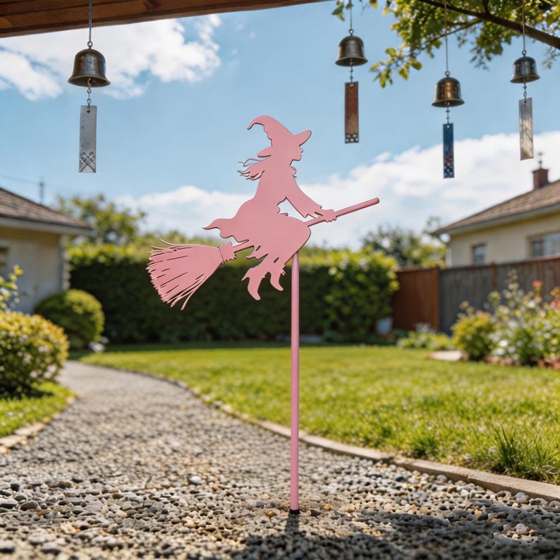 May include: A pink metal witch silhouette on a stake, perfect for garden decor. The witch is riding a broom, with a pointed hat and flowing dress. The background includes a gravel path, green grass, and wind chimes.