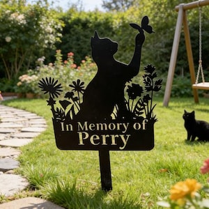 May include: Black metal garden memorial stake featuring a cat silhouette reaching for a butterfly. The stake is inscribed with "In Memory of Perry" and is surrounded by stylized flowers. A black cat sits nearby.