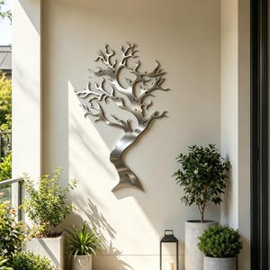 Op de afbeelding: Een zilverkleurige metalen wandsculptuur in de vorm van een gestileerde boom met een gebogen stam en complexe takken. Het object heeft een gladde, geborstelde metalen afwerking en is plat gemonteerd tegen een lichtgekleurde muur.