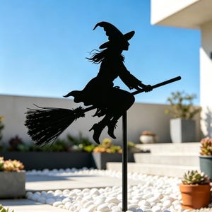 May include: Black metal silhouette of a witch riding a broomstick. The witch is wearing a pointed hat and a flowing dress. The broomstick is detailed with bristles. The artwork is mounted on a black metal stake, suitable for outdoor display.