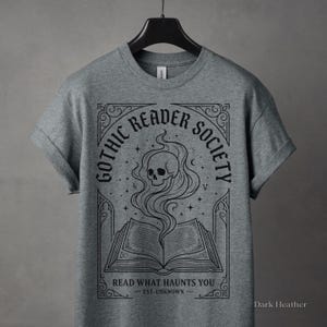 May include: Dark heather gray t-shirt featuring a graphic design for the "Gothic Reader Society." The design includes a skull with smoke rising from an open book, surrounded by text that reads "Read What Haunts You."