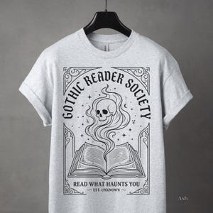 May include: Light gray t-shirt featuring a black graphic with the text "Gothic Reader Society." The design includes an open book with a skull and smoke rising from it. The phrase "Read What Haunts You" is below the book.