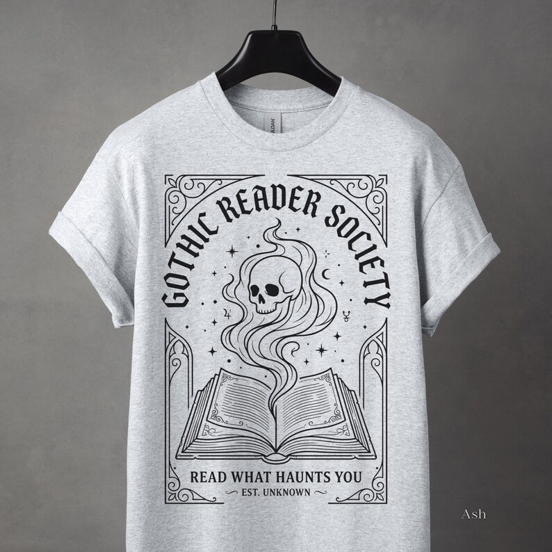 May include: Light gray t-shirt featuring a black graphic with the text "Gothic Reader Society." The design includes an open book with a skull and smoke rising from it. The phrase "Read What Haunts You" is below the book.