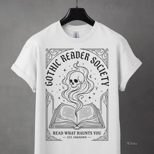 May include: White t-shirt featuring a black graphic design for the "Gothic Reader Society." The design includes an open book with a skull and smoke rising from it, surrounded by text and decorative elements. The text reads "Read What Haunts You."