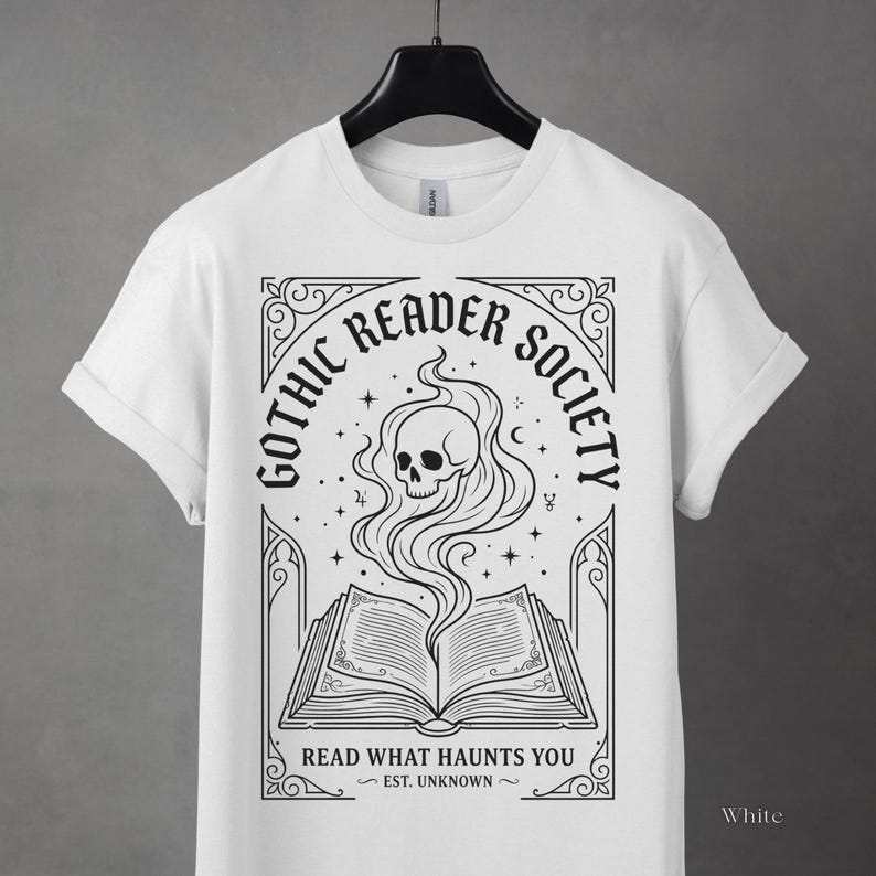 May include: White t-shirt featuring a black graphic design for the "Gothic Reader Society." The design includes an open book with a skull and smoke rising from it, surrounded by text and decorative elements. The text reads "Read What Haunts You."