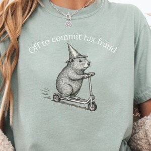 Può includere: T-shirt verde salvia con la scritta "Off to commit tax fraud". La maglietta presenta una grafica di un roditore con un cappello da mago su uno scooter. La maglietta è realizzata in materiale morbido.