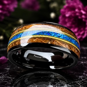 May include: A black ring featuring a central inlay of brown wood and a vibrant blue crushed opal stripe, bordered by two thin gold-toned metallic wires. The interior of the band is polished black and engraved with the text Forever Mine.